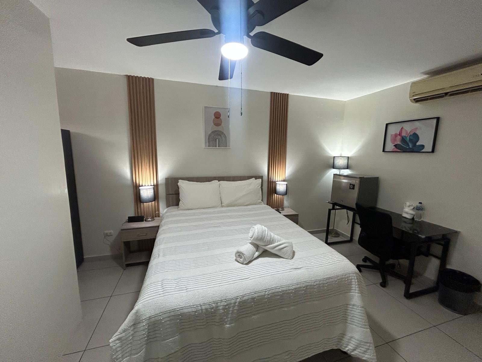 A hotel room with a bed, nightstands, a desk, a chair, a ceiling fan, and an air conditioning unit.