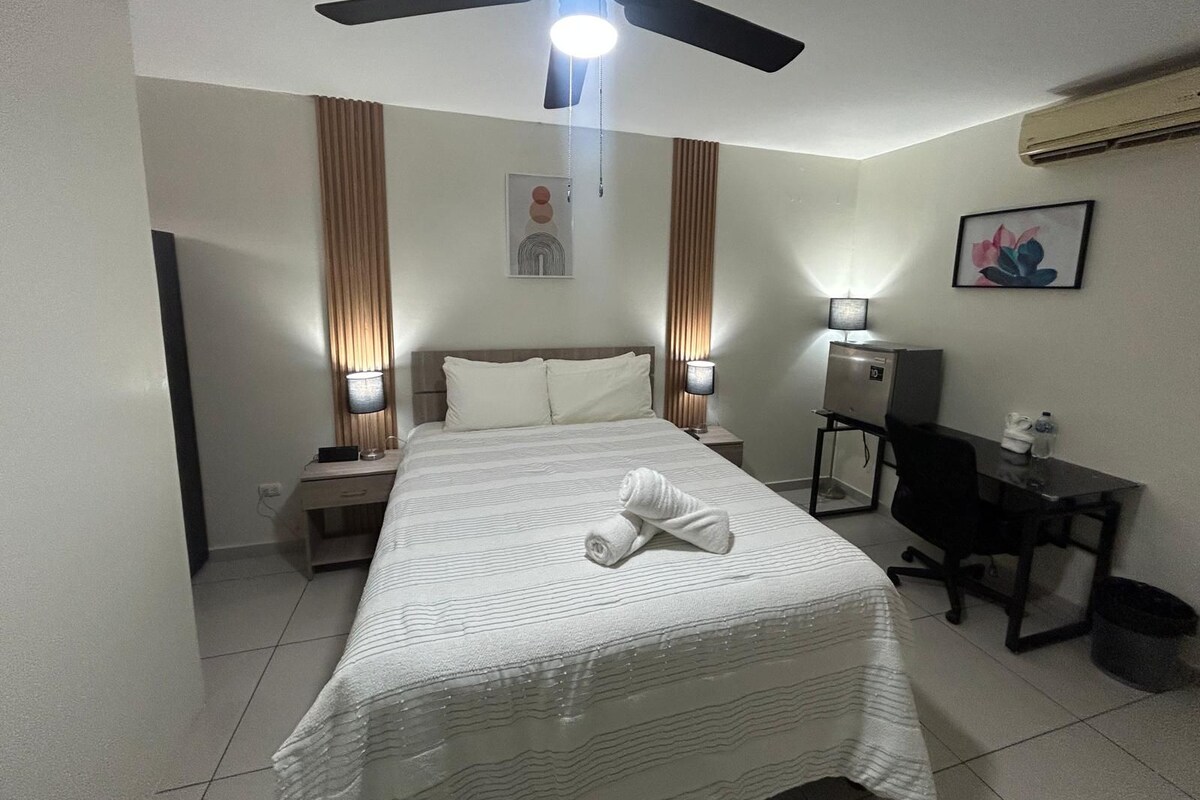 A hotel room with a bed, nightstands, a desk, a chair, a ceiling fan, and an air conditioning unit.