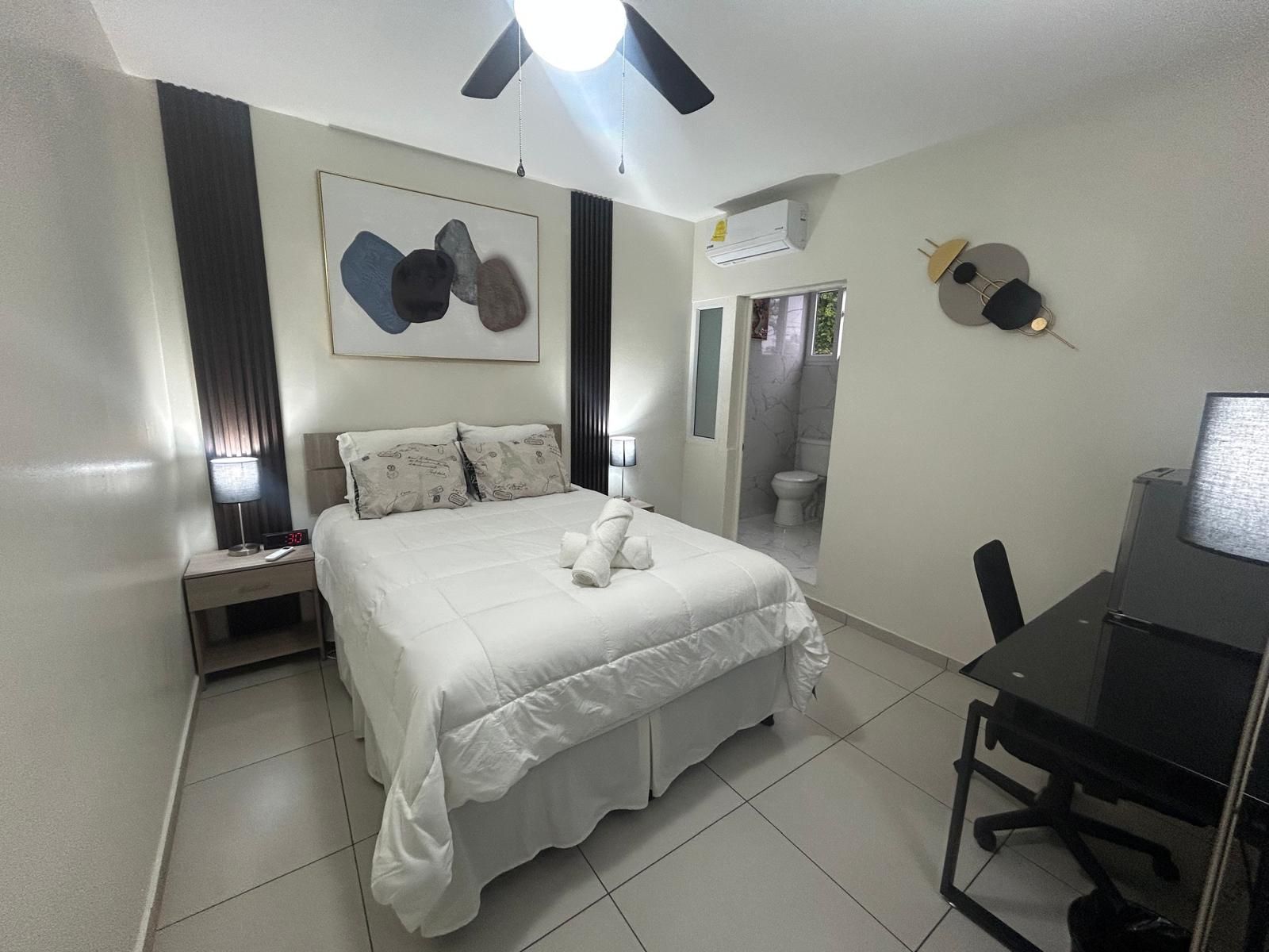 A hotel room with a bed, bedside table, desk, chair, and a bathroom visible through an open door.