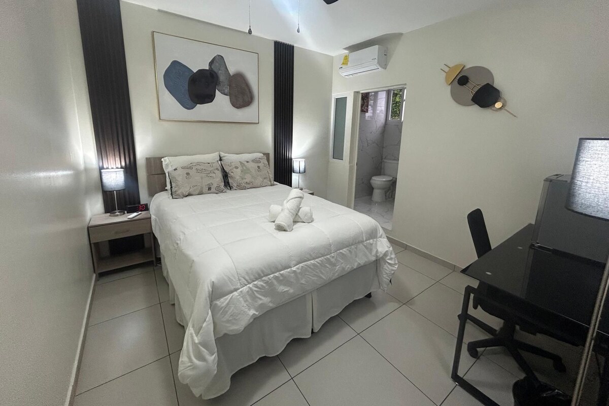 Habitación de hotel con cama, mesita de noche, escritorio, silla y baño visible a través de una puerta abierta.