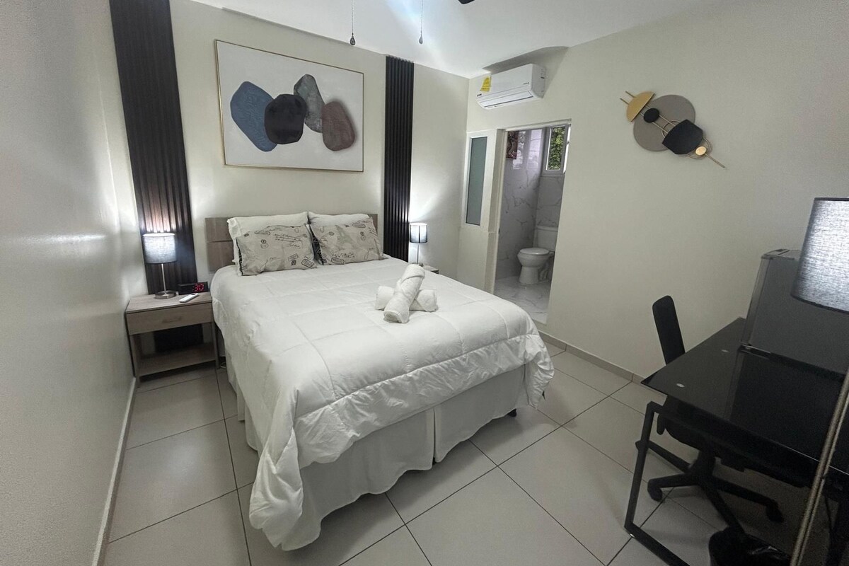 A hotel room with a bed, bedside table, desk, chair, and a bathroom visible through an open door.