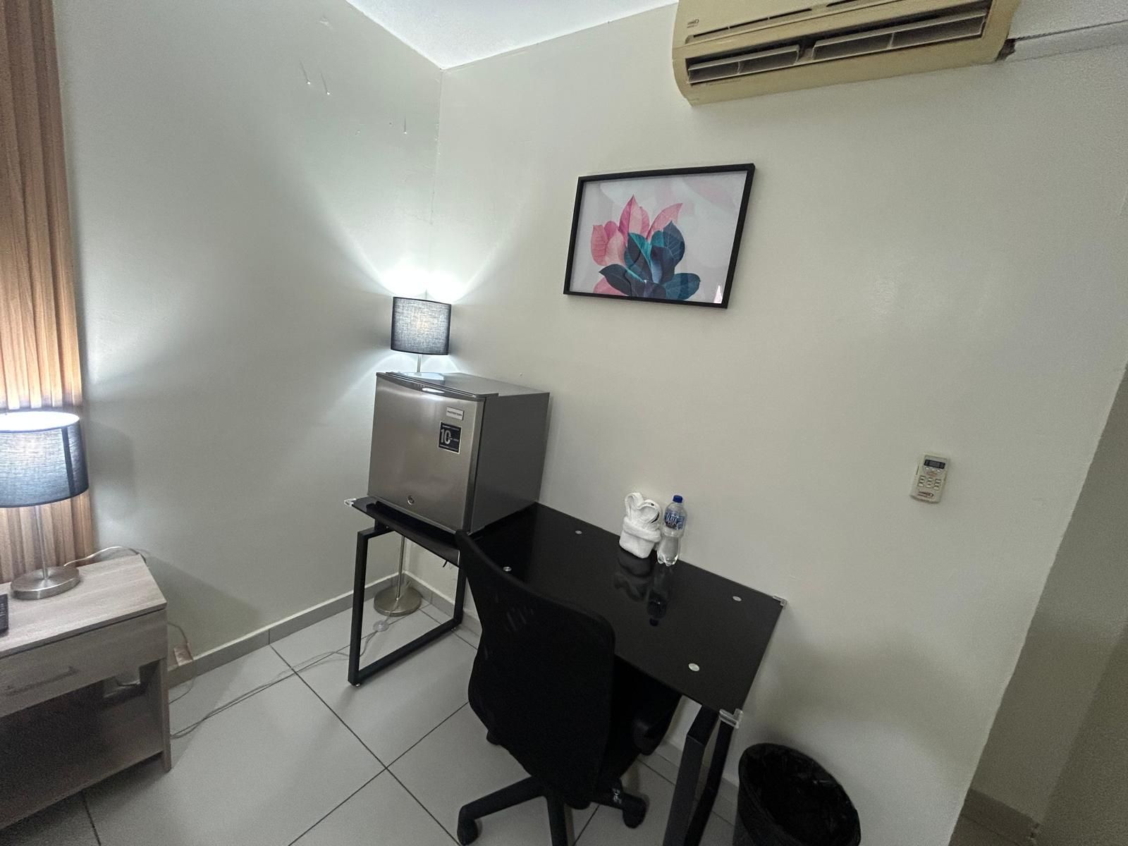 A small office space with a desk, chair, a lamp, a water bottle, a trash bin, and a framed picture on the wall.