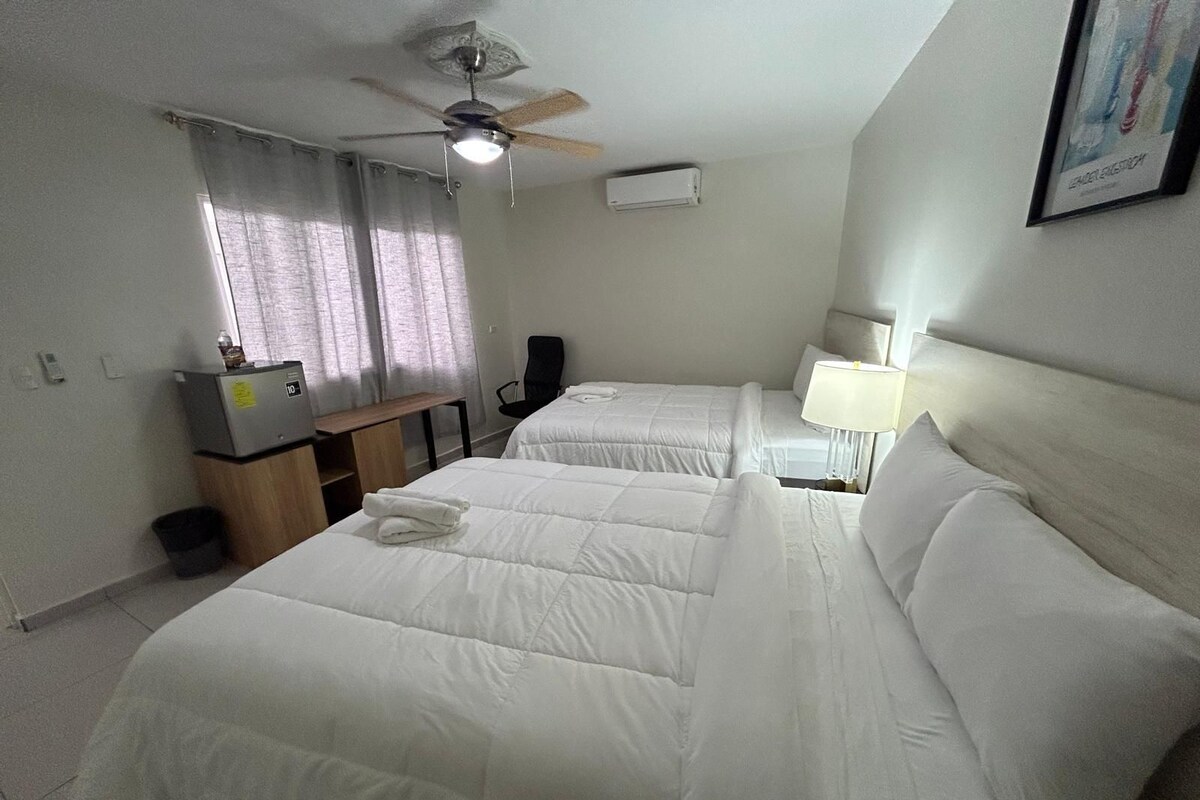 A hotel room with two beds, a desk, a chair, a ceiling fan, and an air conditioning unit.