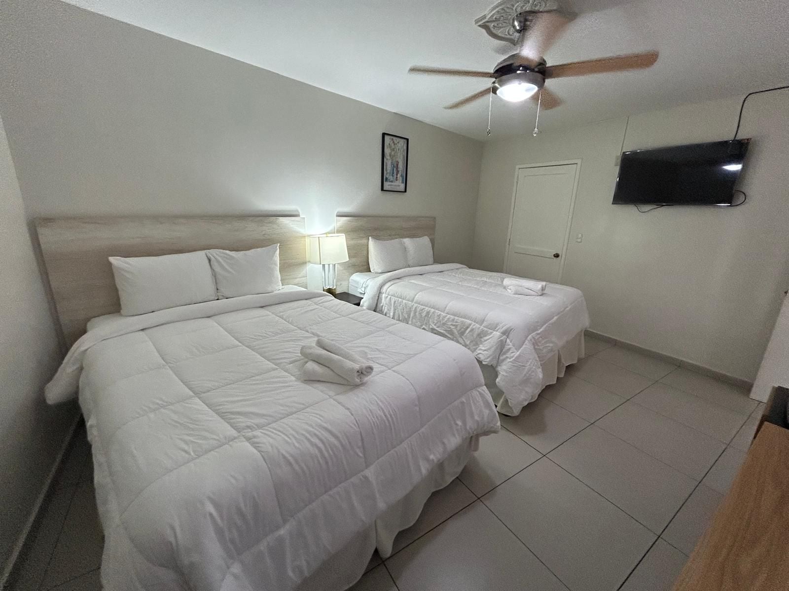 A hotel room with two beds, a ceiling fan, a TV mounted on the wall, and a framed picture on the wall.