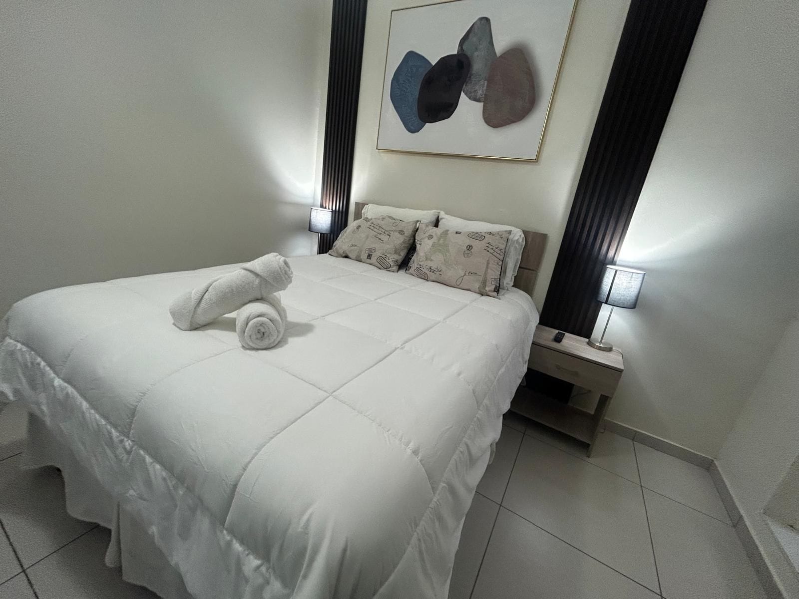 Habitación de hotel con una cama bien hecha, una mesita de noche con lámpara y una obra de arte en la pared.