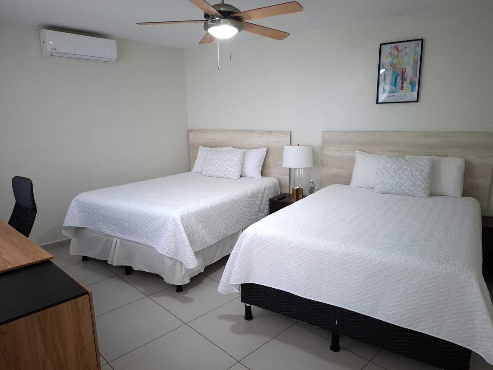 Hotelkamer met twee bedden, een plafondventilator, een airconditioner, een bureau en een ingelijste foto aan de muur.