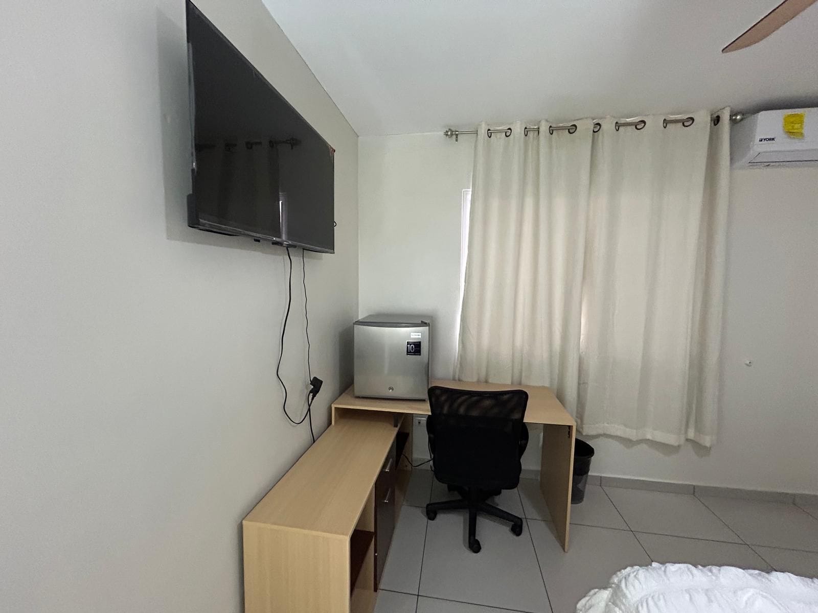 A small, functional room with a bed, a desk, a chair, a television mounted on the wall, and a window with curtains.