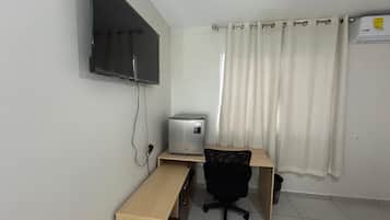 A small, functional room with a bed, a desk, a chair, a television mounted on the wall, and a window with curtains.