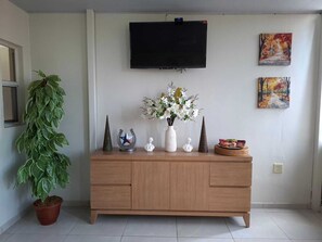 Un salón con un televisor montado en la pared, un mueble de madera con adornos, una planta en maceta y dos cuadros.