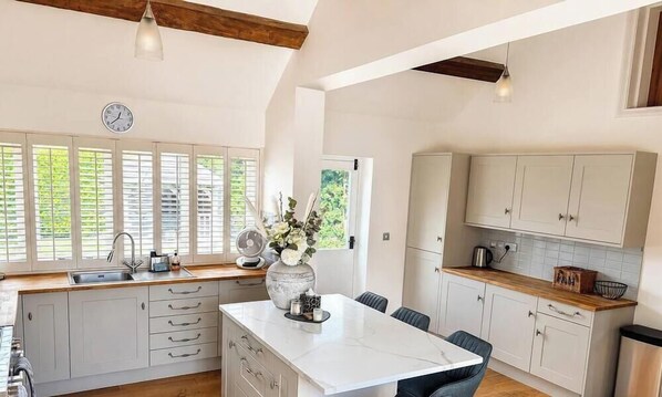 A kitchen with a central island, white cabinetry, a clock, and a window with blinds. - The Old Cowshed - with free parking! (Marchington)