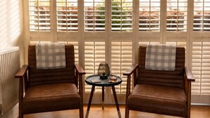 Twee bruine leren fauteuils met gewatteerde kussens met ruitpatroon, een klein rond tafeltje met een lamp en een glas water, en een uitzicht door witte jaloezieën.