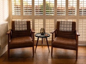 Two brown leather armchairs with checkered throw pillows, a small round table with a lamp and a glass of water, and a view through white blinds. - The Old Cowshed - with free parking! (Marchington)