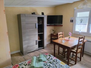 Quarto com armário de madeira, televisão fixada na parede, mesa de jantar com cadeiras e janela com cortina.