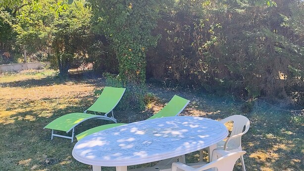 Uma área de estar ao ar livre com uma mesa redonda e quatro cadeiras, cercada por grama e árvores.