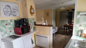 Cozinha compacta com micro-ondas, máquina de café e pequena geladeira. Há uma área de jantar com mesa e cadeiras, e uma cama com colcha florida.