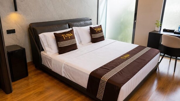 Een moderne hotelkamer met een groot bed, twee kussens met de tekst 'TUNA' geborduurd erop, een nachtkastje en een stoel.