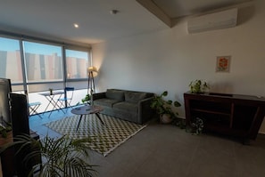 Ein modernes Wohnzimmer mit einer Couch, einem kleinen runden Tisch, einer Stehlampe und einem Fernseher. Es gibt einen Balkon mit Blick auf Gebäude.