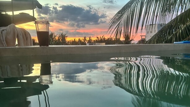 Une piscine à l’eau claire où se reflètent les palmiers et le ciel au coucher du soleil.
