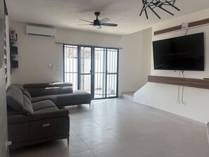 A living room with a sectional sofa, a ceiling fan, a flat-screen TV, and a large window with a view. - Casa 5 minutos del Foro GNP Américas (Mérida)