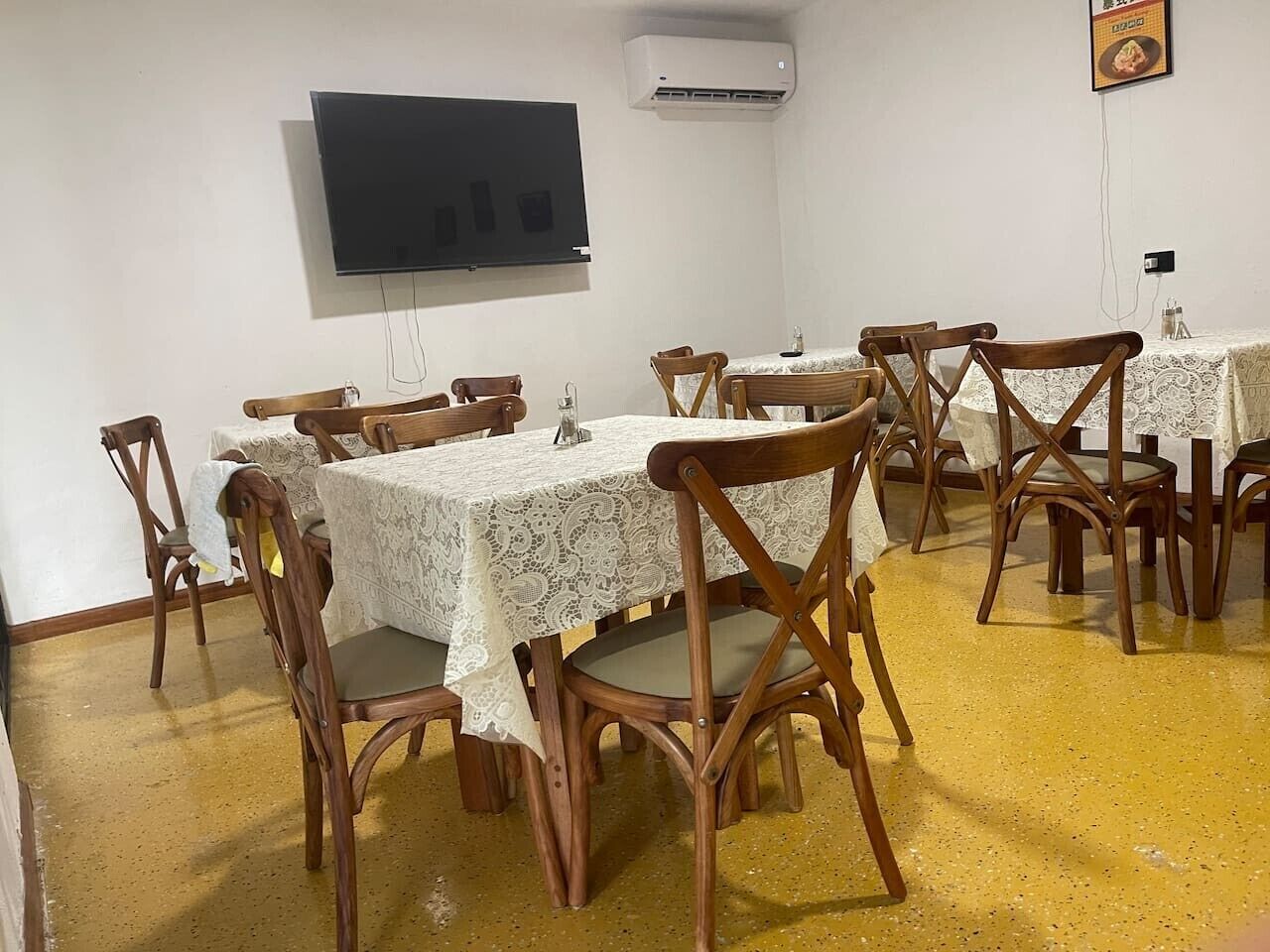 A dining area with tables covered in lace tablecloths, wooden chairs, a flat-screen TV mounted on the wall, and an air conditioning unit.
