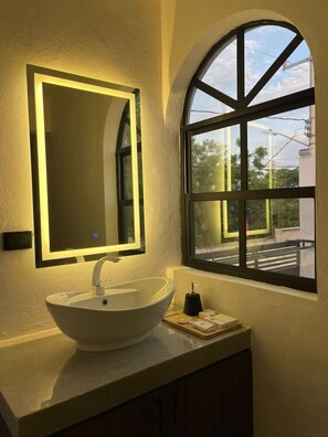 Ein Badezimmer mit einem weißen Waschtisch, einem Spiegel mit indirekter Beleuchtung und einem Fenster mit Blick ins Freie.