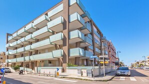 Un edificio de varios pisos con balcones, cielo despejado y una calle con automóviles y un paso de peatones.