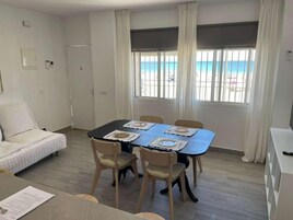 Una habitación con cama, una mesa de comedor con sillas, una ventana con vista a la playa y una puerta.