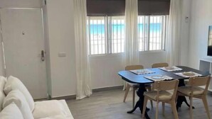 A room with a sofa, a dining table with chairs, a small round ottoman, and a view of the sea through the windows. - Córdoba 8 by 7Escalones (Rota)