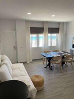 Una habitación con sofá, una mesa de comedor con sillas, un otoman pequeño y redondo, y vista al mar por las ventanas.