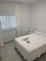 Una habitación con una cama, una mesita de noche, un ventanal con cortinas y suelo de baldosas.