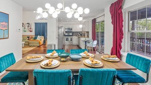 Un comedor con sillas tapizadas en color verde azulado, una mesa de madera dispuesta con platos y servilletas, una araña con varias luces y vistas a la cocina y la sala de estar a través de una puerta abierta.