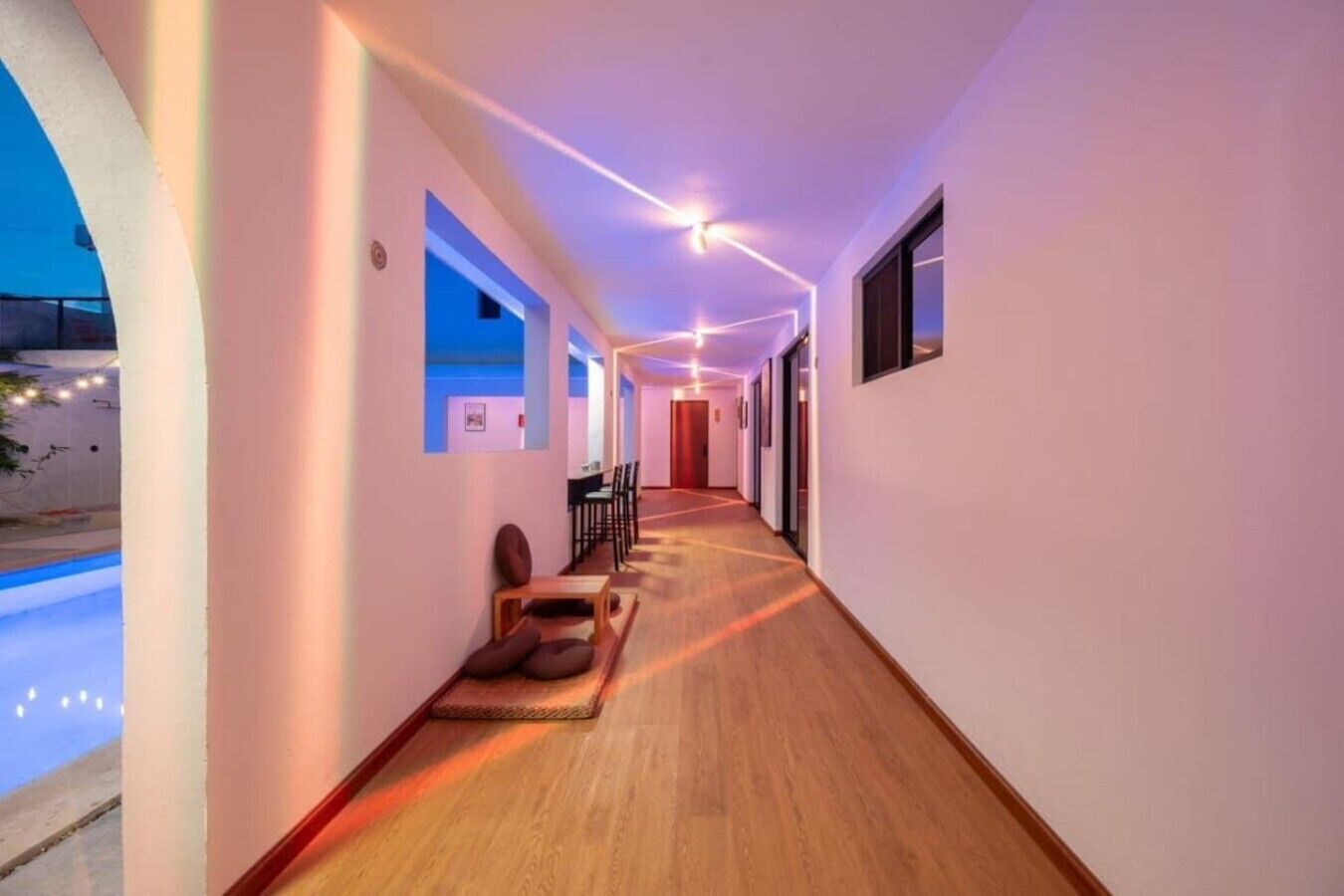 Un corridoio d'albergo con pavimento in legno, un'area relax con cuscini e vista sulla piscina esterna.