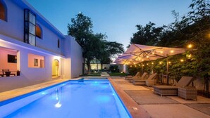 Une piscine dont le bord est illuminé de lumière bleue, avec des chaises longues et des parasols sous des guirlandes lumineuses.