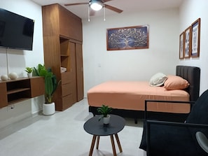 Un espace de vie compact comprenant un lit, un téléviseur, un ventilateur de plafond, une petite table et une plante en pot.