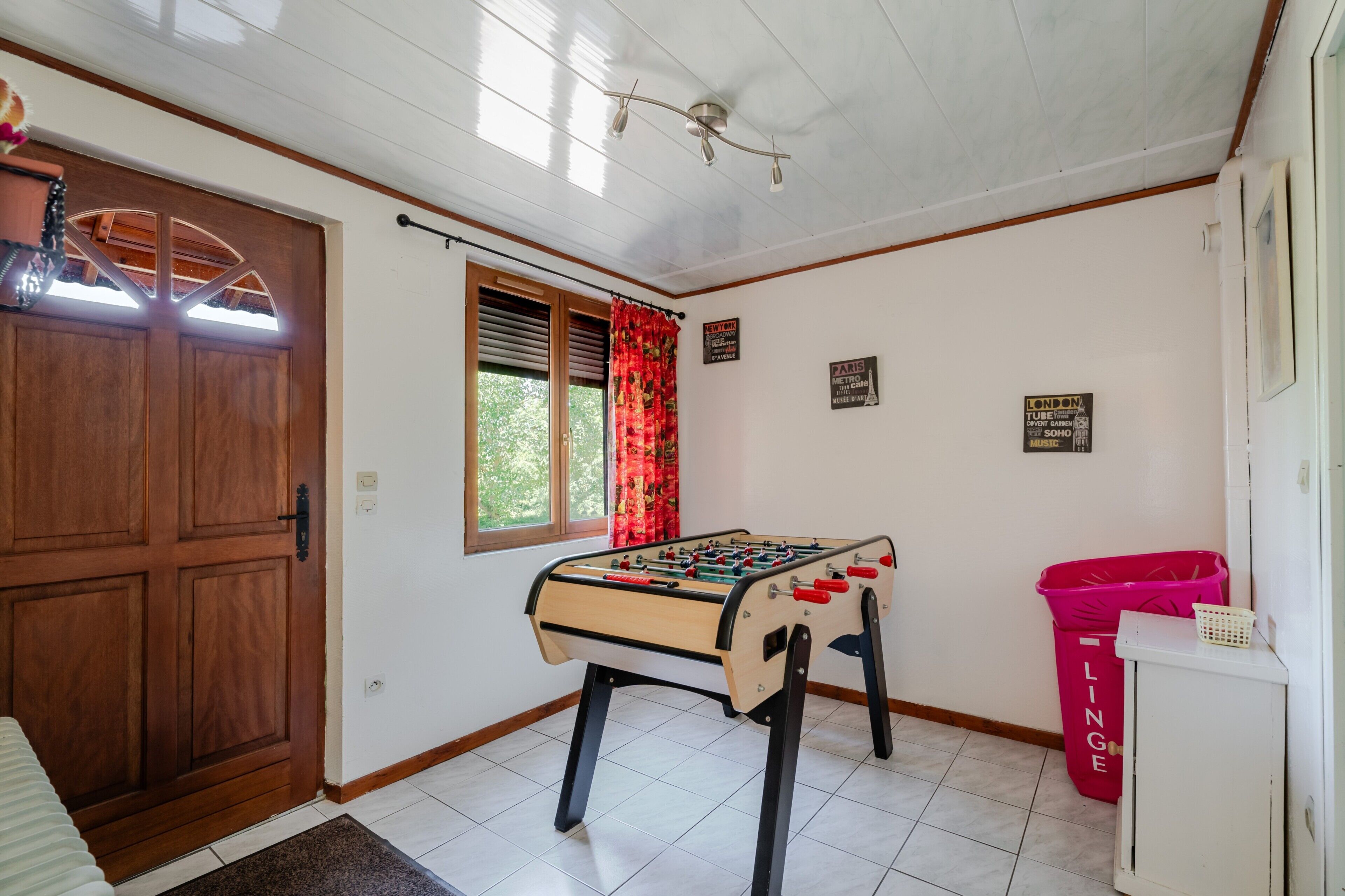 Um quarto com porta de madeira, janela com cortinas vermelhas, mesa de futebol, lixeira e decorações nas paredes.