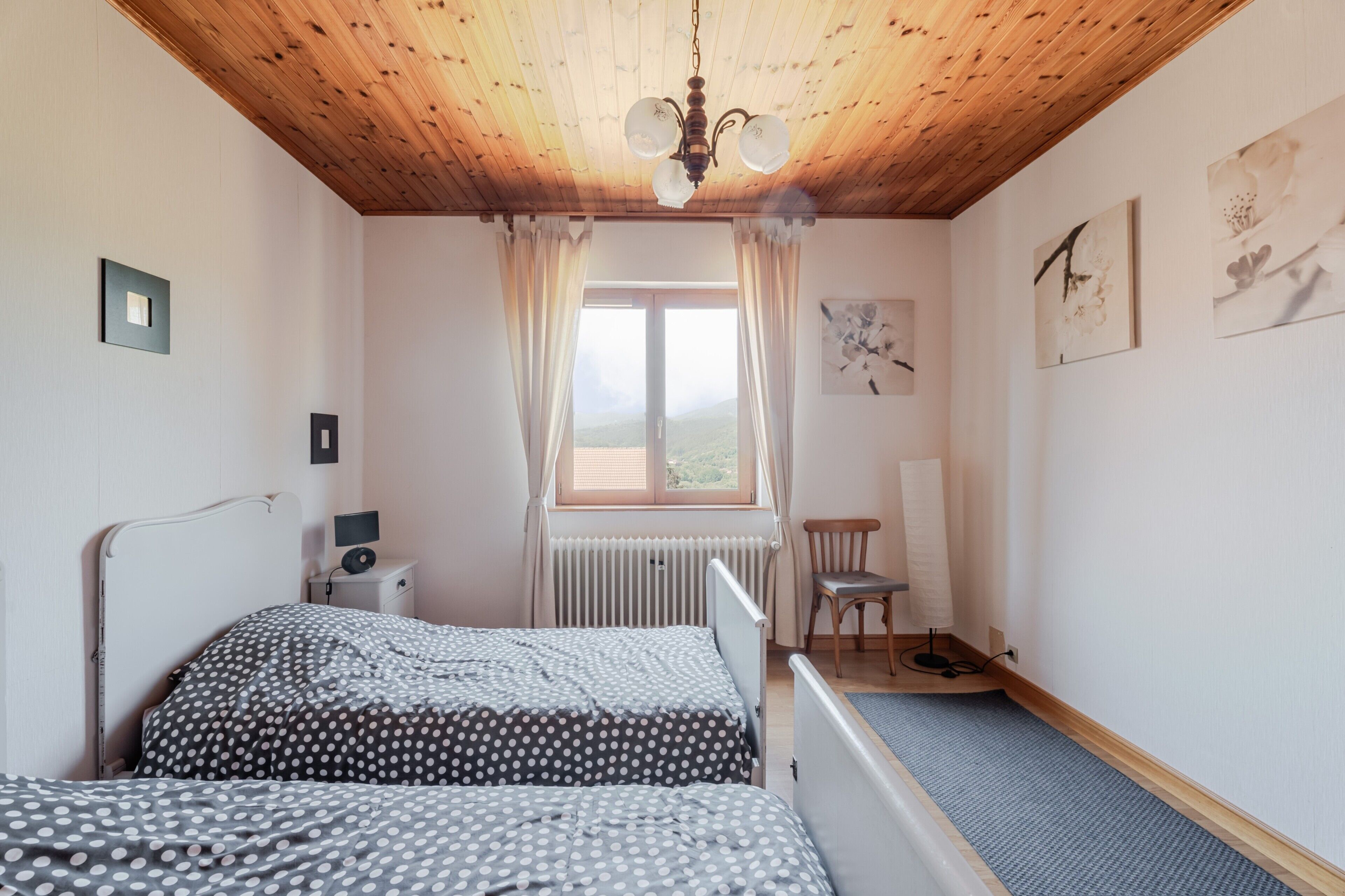A bedroom with two beds, a wooden ceiling, a window with curtains, a radiator, and a chair.