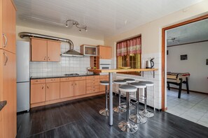 A modern kitchen with wooden cabinets, a central island with bar stools, a window with checkered curtains, and a visible radiator. - Gite de la Bruche in Schwartzbach (Russ)