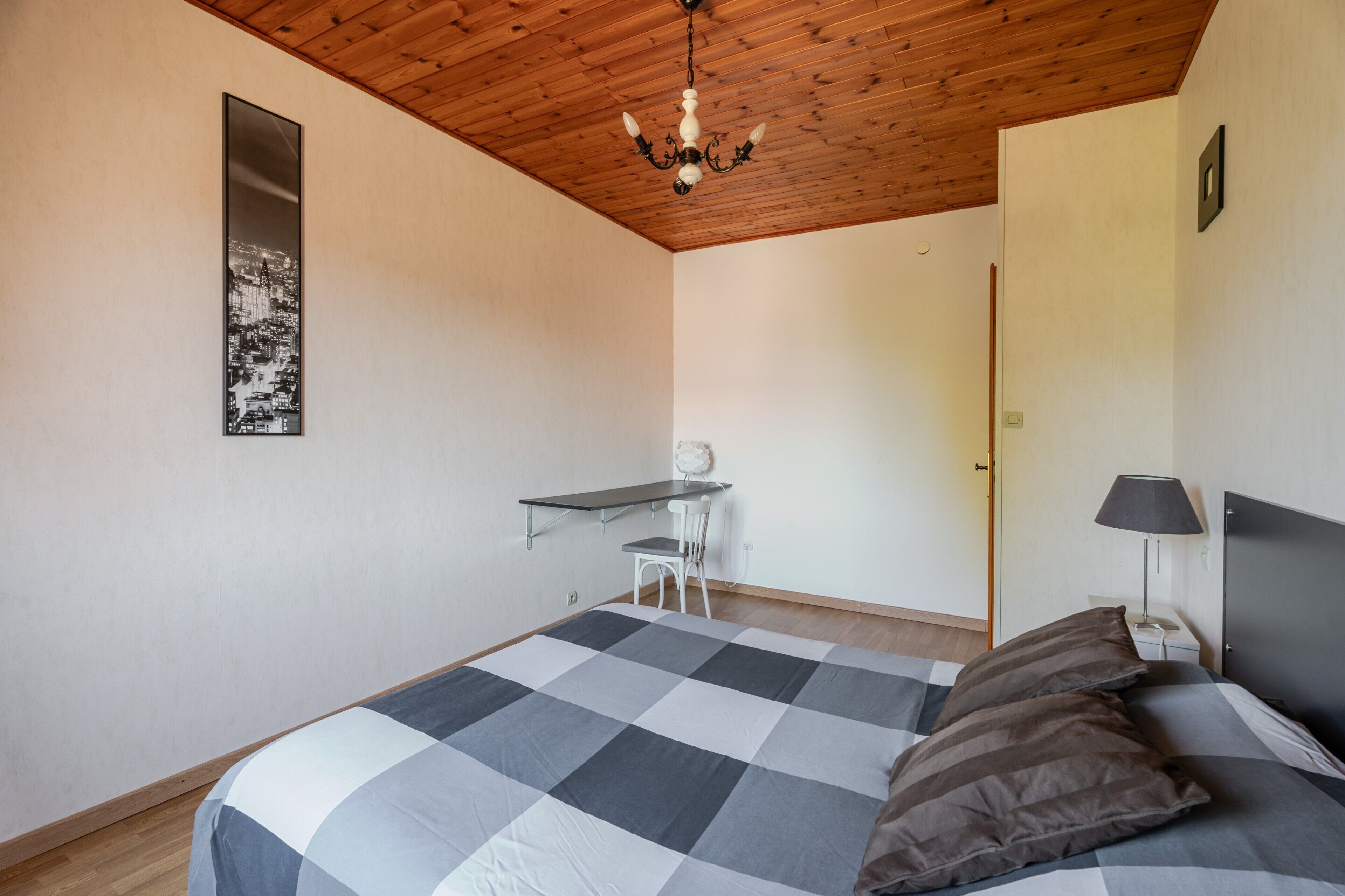 A bedroom with a bed, a desk, a chair, a lamp, and a cityscape artwork on the wall.