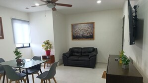 Una sala de estar con un sofá, una mesa de comedor con sillas, un ventilador de techo, un cuadro en la pared y un televisor montado en la pared.