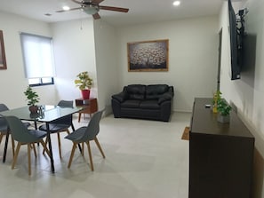 Un soggiorno con un divano, un tavolo da pranzo con sedie, una ventola a soffitto, un quadro appeso al muro e una TV fissata alla parete.