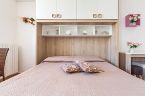 Une chambre à coucher avec un lit, des oreillers, une commode à étagÚres ouvertes, une chaise et une petite table sur laquelle est posé un vase.