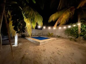 Ein sandiger Außenbereich mit einem blauen Pool, Palmen und Lichterketten.