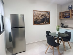 Ett kompakt kök med ett kylskåp, ett matbord med stolar, en liten krukväxt och en tavla på väggen.