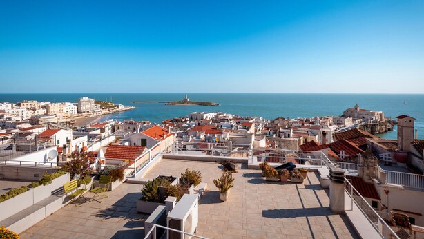Un paisaje urbano costero con vista al mar, edificios con techos de tejas rojas y un cielo despejado.