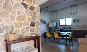 Un mur en pierre, une table en bois avec un récipient en verre, un coin repas avec des chaises jaunes, une cuisine équipée d’un four à micro-ondes et une fenêtre laissant entrer la lumière du jour.