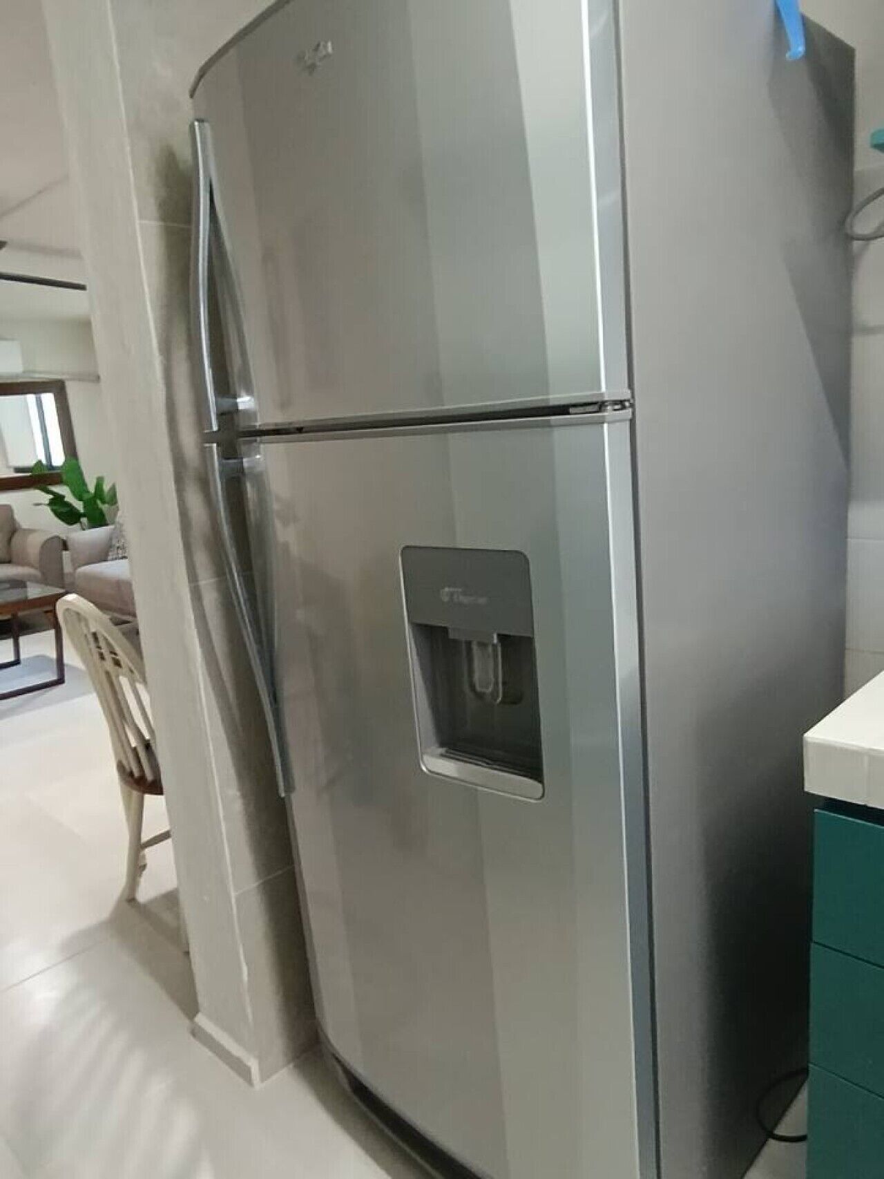 A stainless steel refrigerator with a water dispenser, located in a modern kitchen with white cabinetry and a dining area visible in the background.