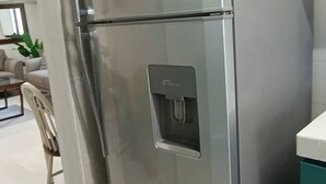 Un refrigerador de acero inoxidable con dispensador de agua, situado en una cocina moderna con armarios blancos y un comedor visible al fondo.