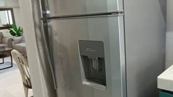 A stainless steel refrigerator with a water dispenser, located in a modern kitchen with white cabinetry and a dining area visible in the background.
