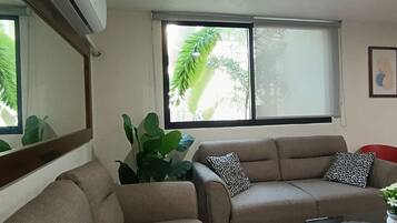 A living room with a beige sofa, a red chair, a large potted plant, and a window with blinds.