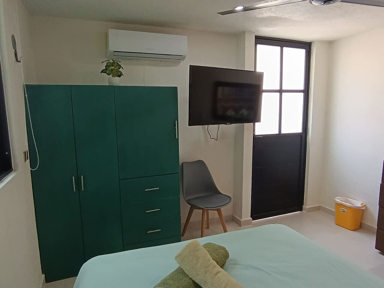 A bedroom with a bed, a green wardrobe, a chair, a TV, and a window.
